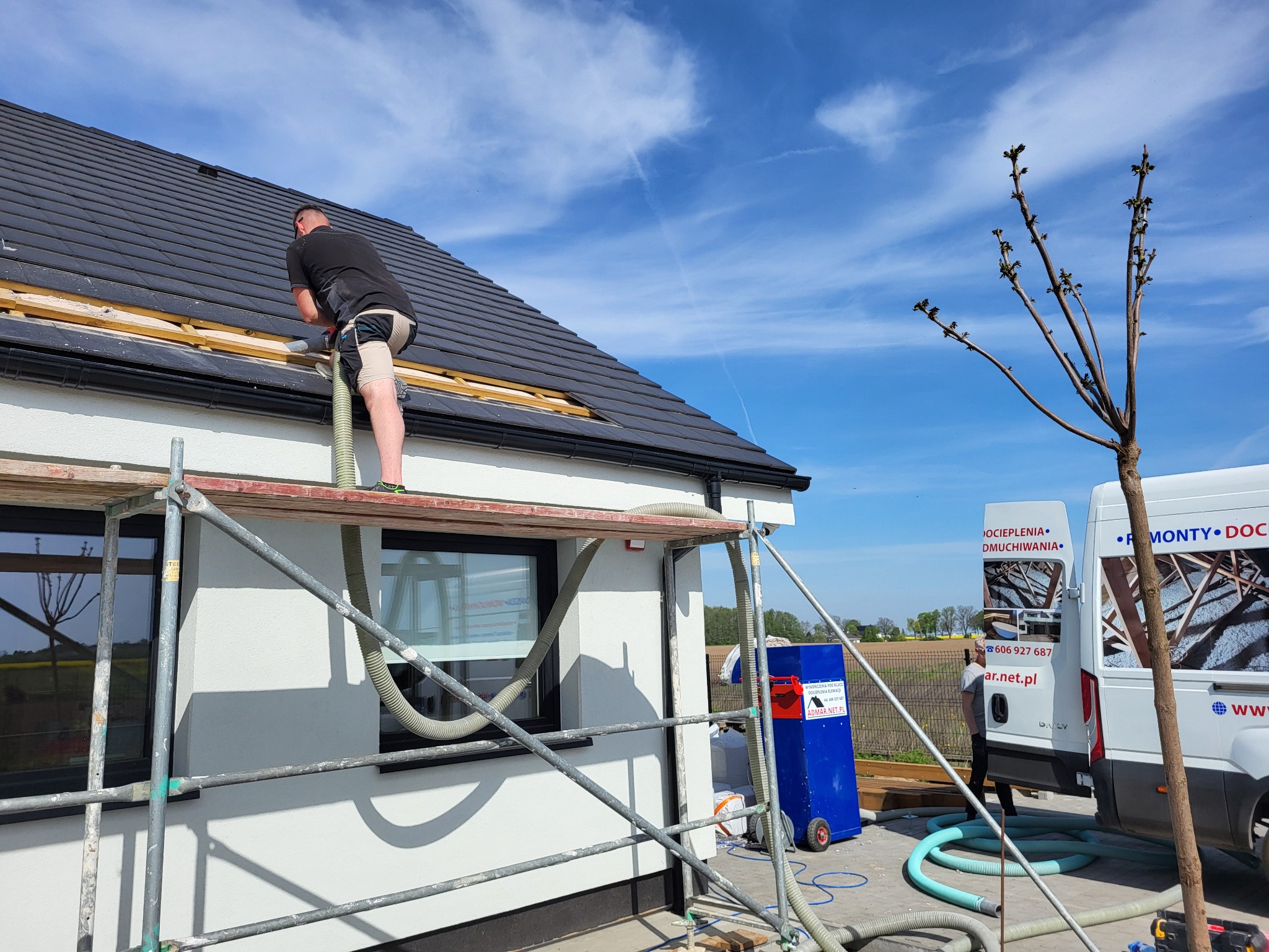 Pracownik na rusztowaniu aplikuje materiał izolacyjny w szczelinę dachu domu jednorodzinnego, widoczna maszyna do wdmuchiwania izolacji oraz firmowy samochód dostawczy