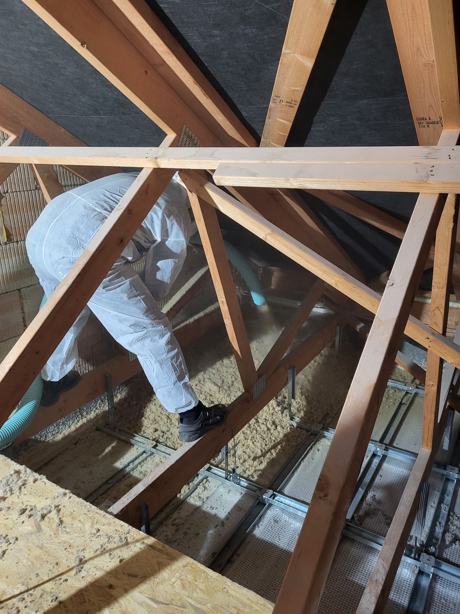 Pracownik w kombinezonie ochronnym aplikuje materiał izolacyjny na poddaszu za pomocą węża, widoczne drewniane elementy konstrukcji dachu oraz metalowy stelaż pod podłogą.