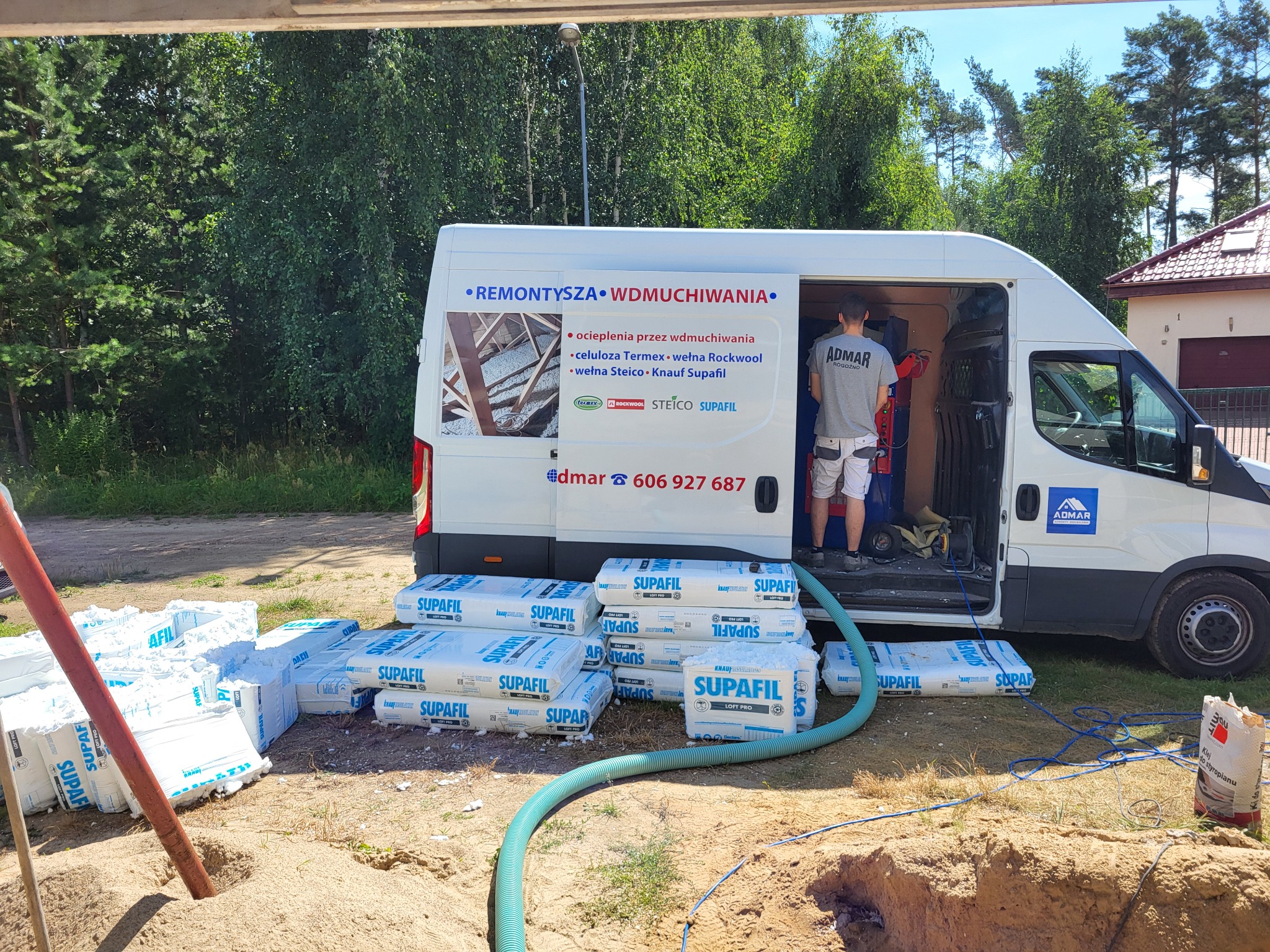 Ocieplanie wdmuchiwaniem: biały bus firmy ADMAR z otwartymi drzwiami, pracownik w środku, na zewnątrz worki z wełną Supafil i wąż do wdmuchiwania, widoczne drzewa i dom w tle.