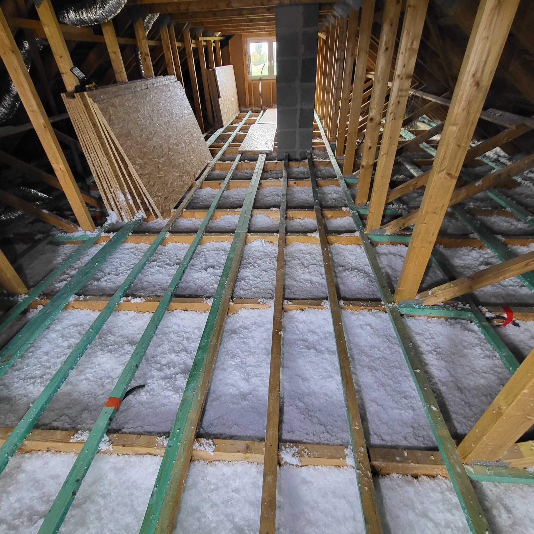 Poddasze ocieplone białą wełną mineralną, widoczne drewniane krokwie i zielone legary, w tle komin i okno, na lewo stos płyt OSB.