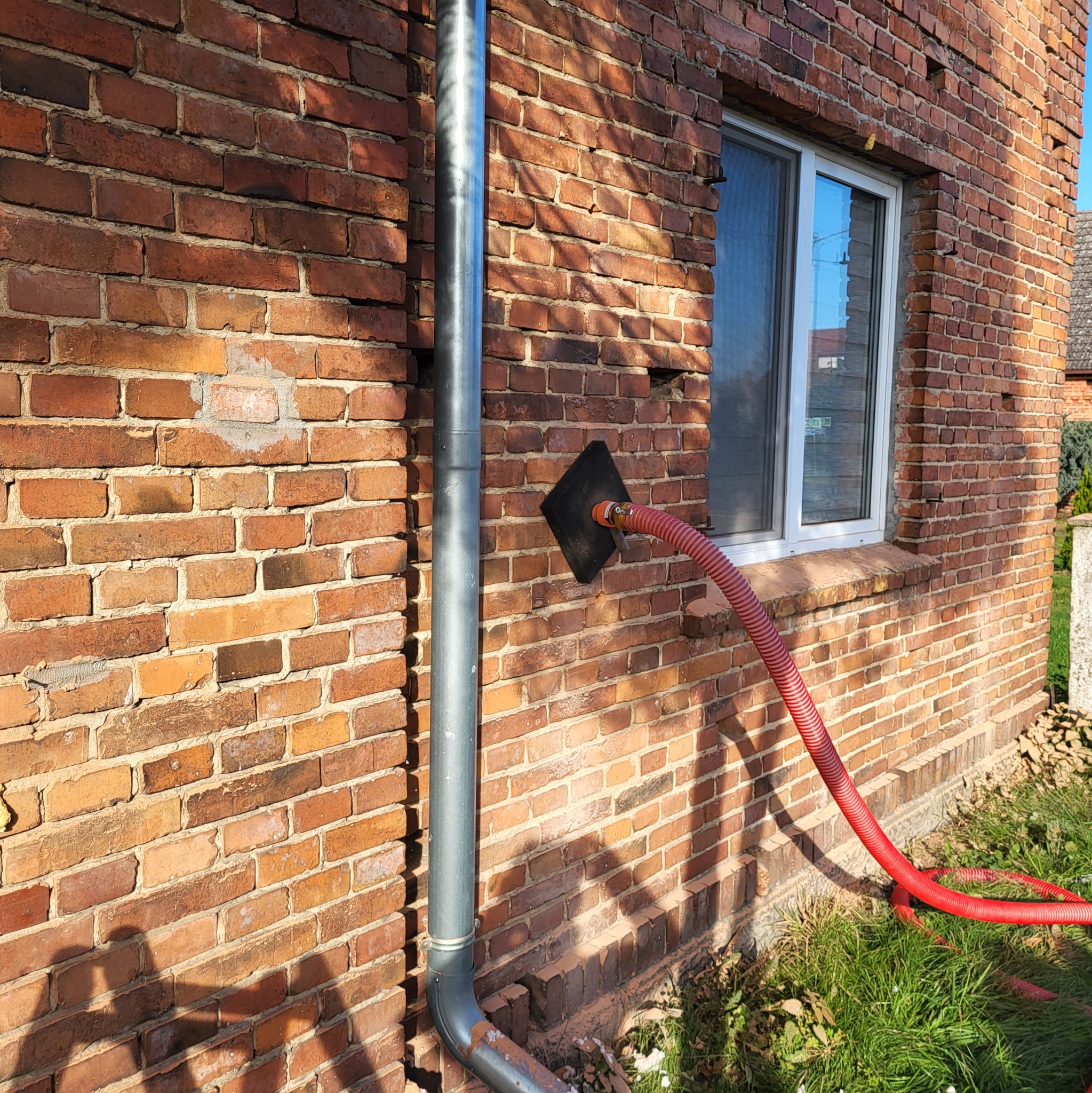 Czerwony wąż przymocowany do czarnej płyty na ceglanej ścianie budynku, obok rynny, prawdopodobnie podczas prac związanych z ociepleniem.