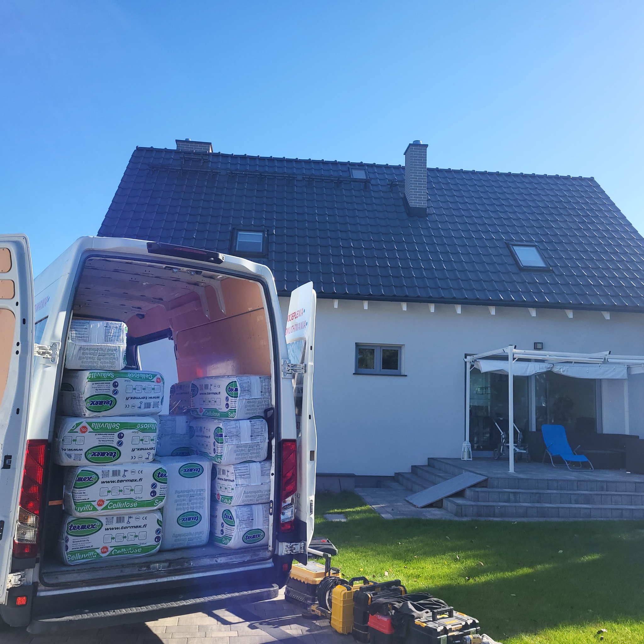 Van wypełniony paczkami izolacji celulozowej Termex Selluvilla przed domem z ciemną dachówką i jasną elewacją, na trawniku leżą narzędzia.