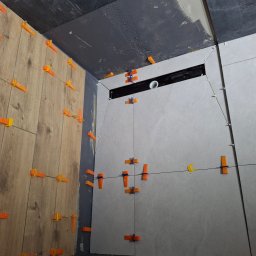 Dawid Adamczuk - Układanie płytek drewnopodobnych i szarych w łazience z użyciem klipsów poziomujących i sznurka, widoczny odpływ liniowy w ścianie.