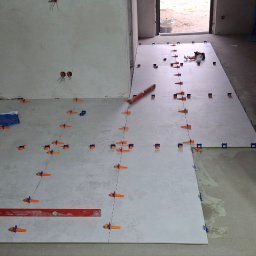 Dawid Adamczuk - Układanie jasnych płytek podłogowych z użyciem poziomicy i pomarańczowych krzyżyków dystansowych w trakcie remontu łazienki, widoczne instalacje wodne i elektryczne.