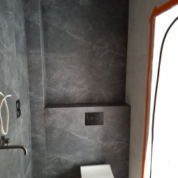 Dawid Adamczuk - Nowoczesna łazienka wykończona płytkami imitującymi kamień w odcieniach szarości, z podtynkową toaletą i minimalistyczną armaturą w kolorze ciemnego chromu. Zabezpieczone folią malarską obramowanie...