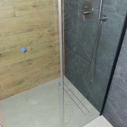 Dawid Adamczuk - Nowoczesna kabina prysznicowa z odpływem liniowym, wykończona płytkami imitującymi drewno i szarym kamieniem, bateria podtynkowa z deszczownicą, widoczna niebieska naklejka informacyjna.