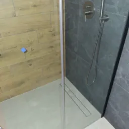 Nowoczesna kabina prysznicowa z odpływem liniowym, wykończona płytkami imitującymi drewno i szarym kamieniem, bateria podtynkowa z deszczownicą, widoczna niebieska naklejka informacyjna.