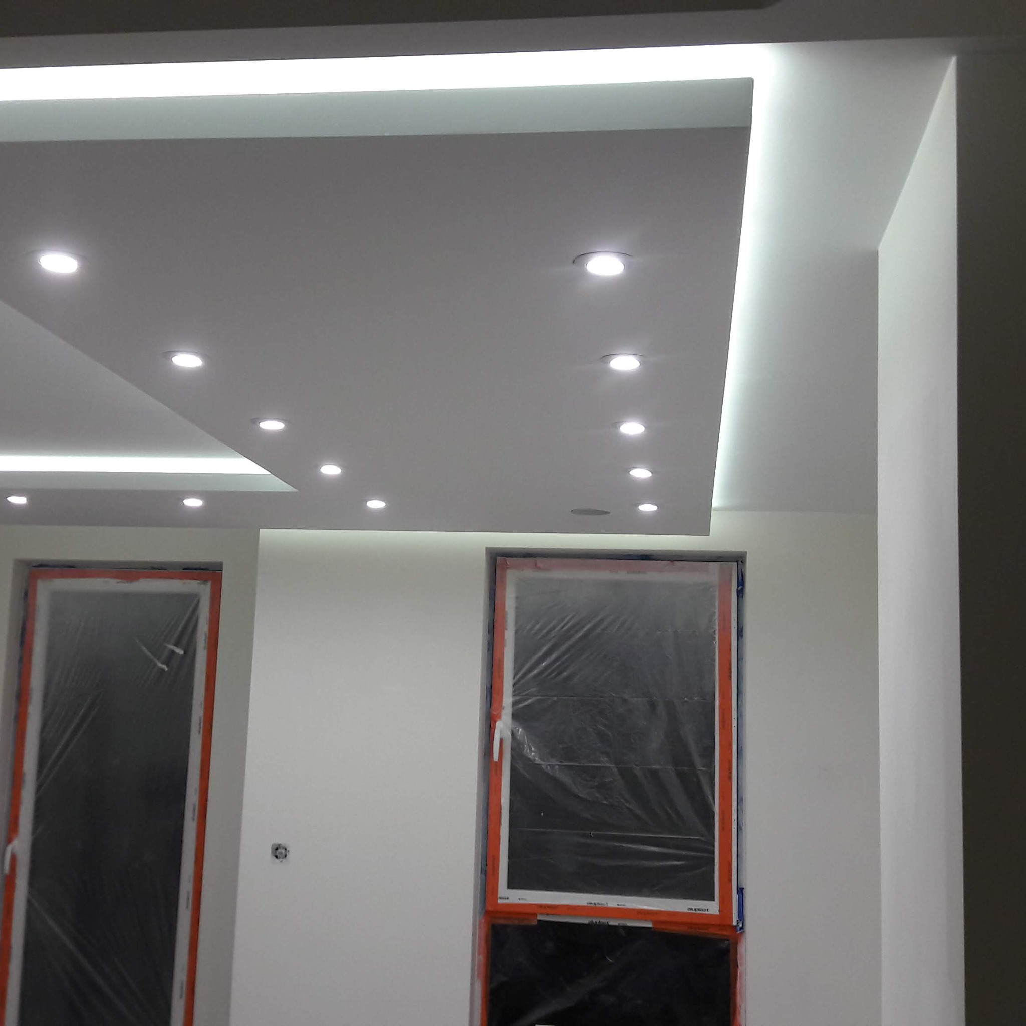 Nowoczesny sufit podwieszany z oświetleniem LED punktowym i listwą LED, okna zabezpieczone folią malarską, ściany przygotowane do malowania.