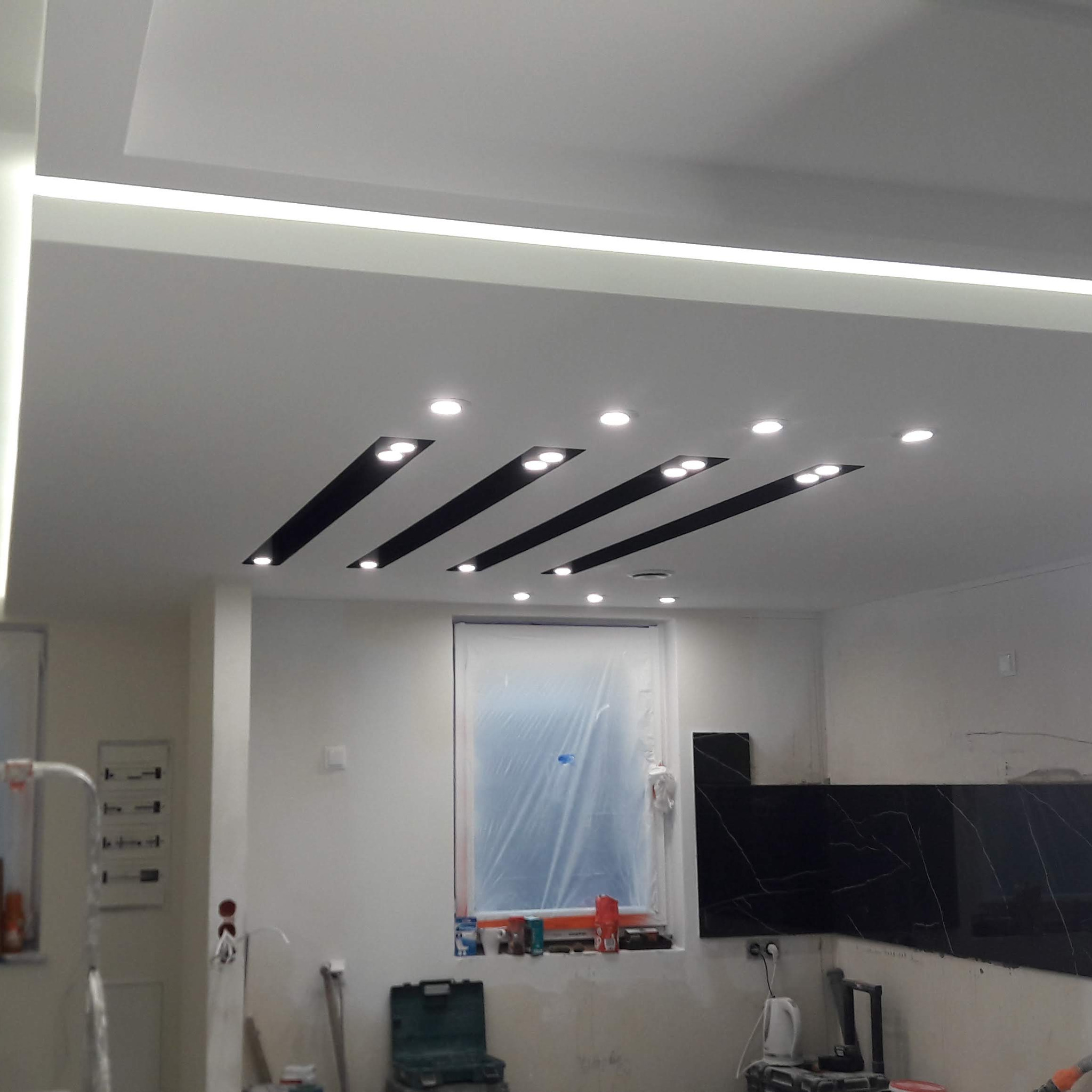 Remontowane wnętrze z podwieszanym sufitem z oświetleniem LED i punktowymi lampami, okno zabezpieczone folią malarską, widoczne narzędzia i materiały budowlane.