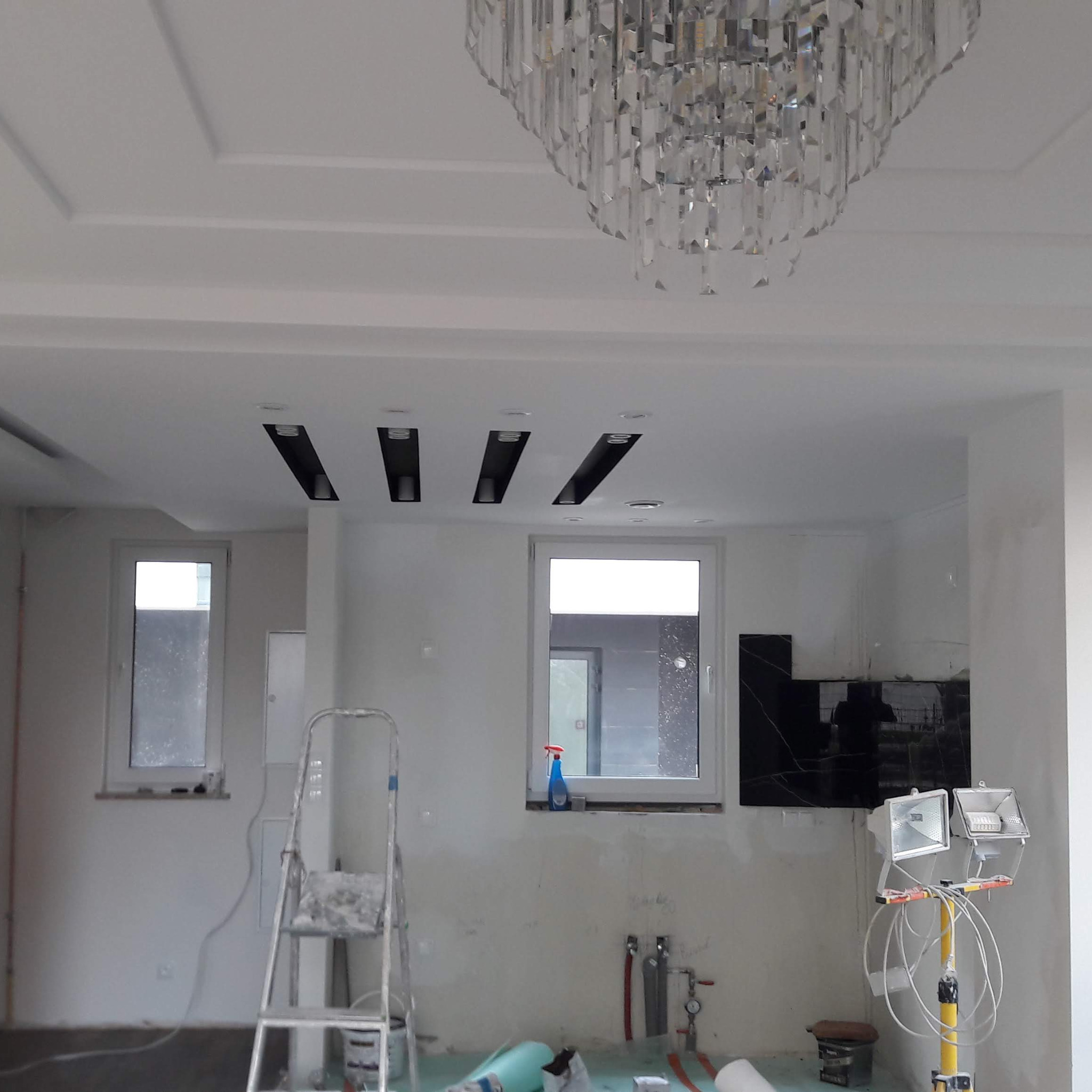 Wnętrze w trakcie prac wykończeniowych: widoczny sufit z ozdobnymi listwami i kryształowym żyrandolem, okna, drabina, reflektory budowlane i fragment ściany z czarną okładziną.