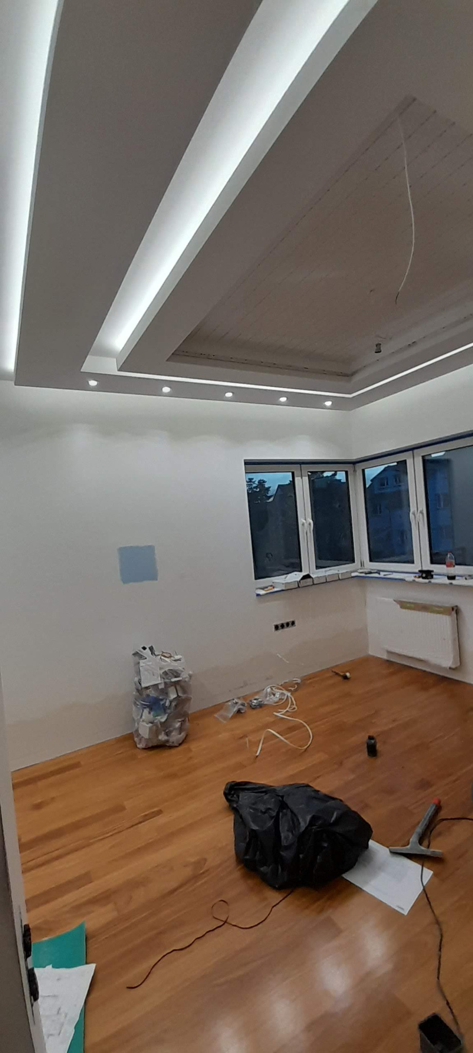 Pokój po częściowym remoncie z drewnianą podłogą, świeżo pomalowanymi ścianami z próbką koloru, ozdobnym podwieszanym sufitem z oświetleniem LED, workiem gruzu i narzędziami na podłodze.