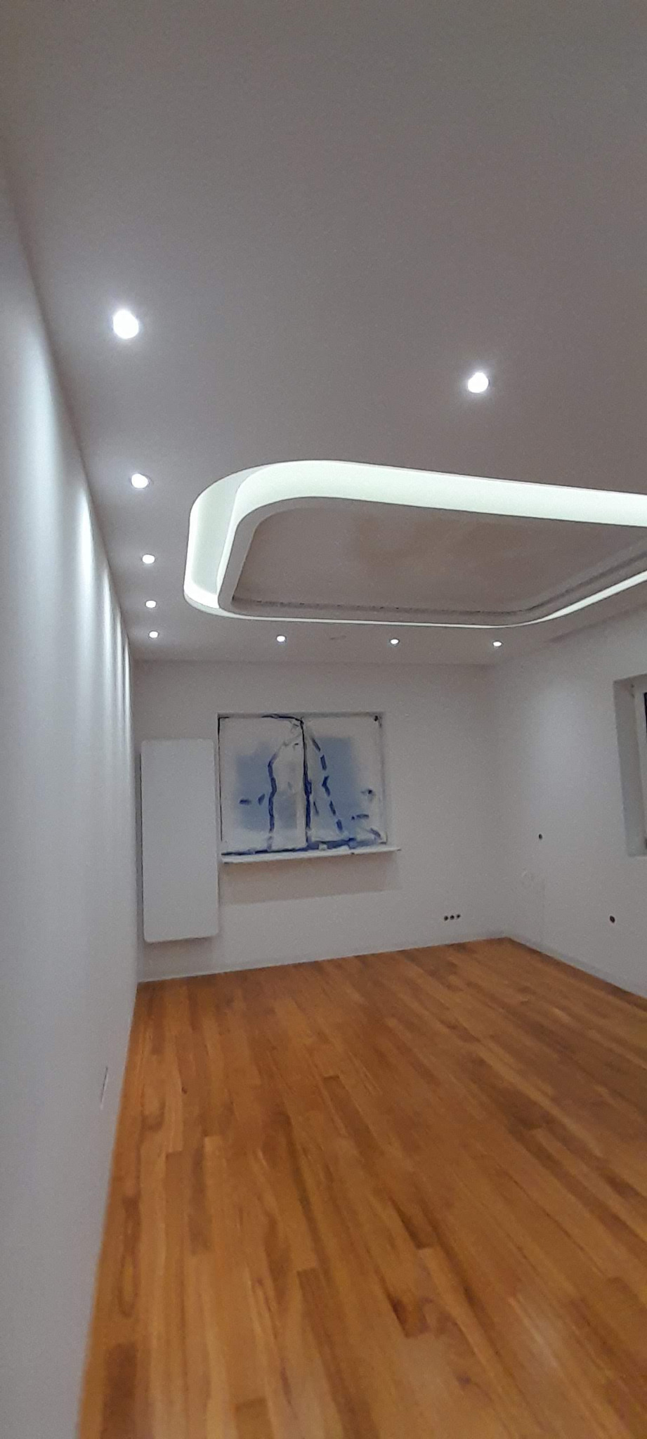 Jasne, przestronne wnętrze po remoncie z drewnianą podłogą, białymi ścianami i podwieszanym sufitem z oświetleniem LED. Okna zabezpieczone folią malarską.