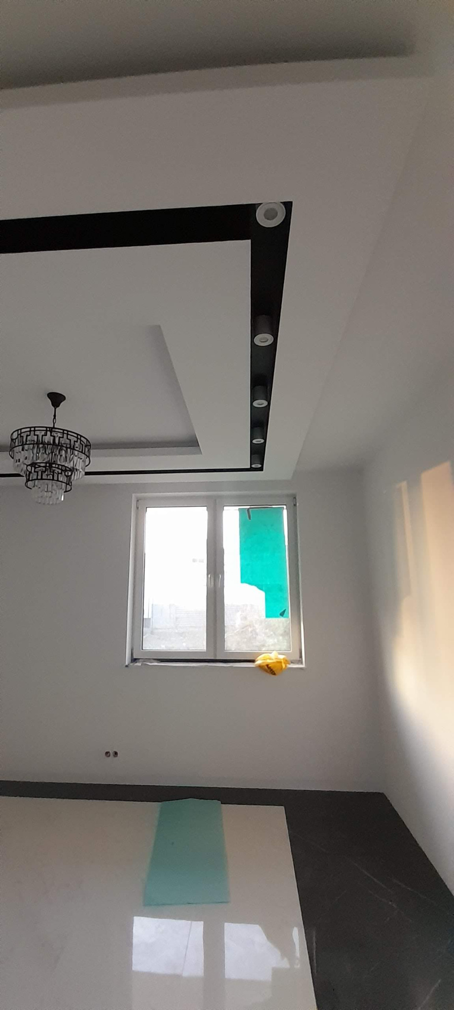Wnętrze pokoju po remoncie: białe ściany, biała podłoga częściowo zakryta folią ochronną, czarny sufit z oświetleniem punktowym i ozdobny żyrandol, okno z folią ochronną.