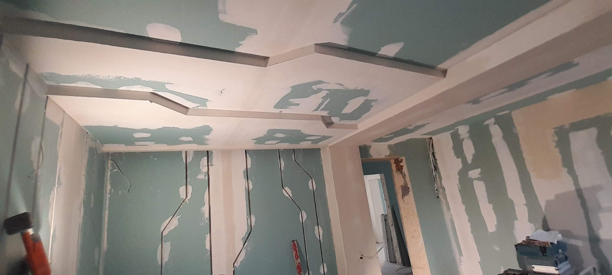 Wnętrze w trakcie prac wykończeniowych: ściany z płyt gipsowo-kartonowych z widocznymi łączeniami, sufit z dekoracyjnymi elementami gipsowymi przed szpachlowaniem, widoczne narzędzia i poziomica.