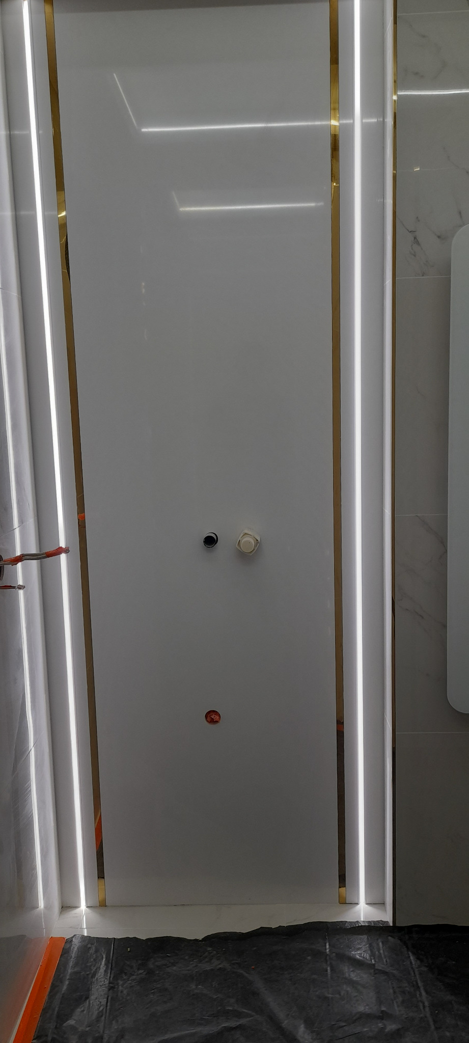 Ściana łazienki z białymi panelami i złotymi akcentami, oświetlenie LED wzdłuż krawędzi, widoczne otwory na instalacje, zabezpieczona podłoga.