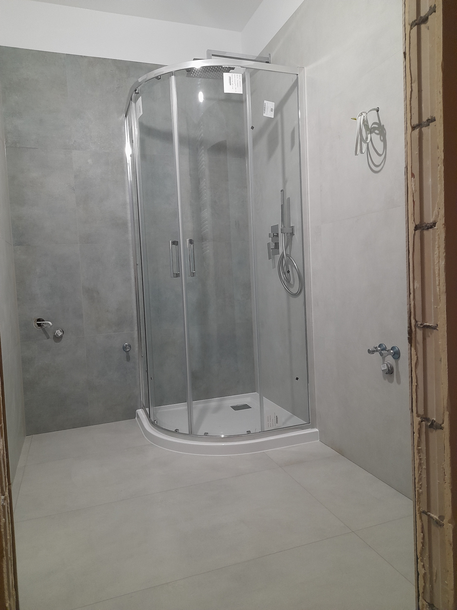 Narożna kabina prysznicowa z chromowanymi detalami, zamontowana w łazience wykończonej szarą płytką, widoczne wystające przewody na ścianie.