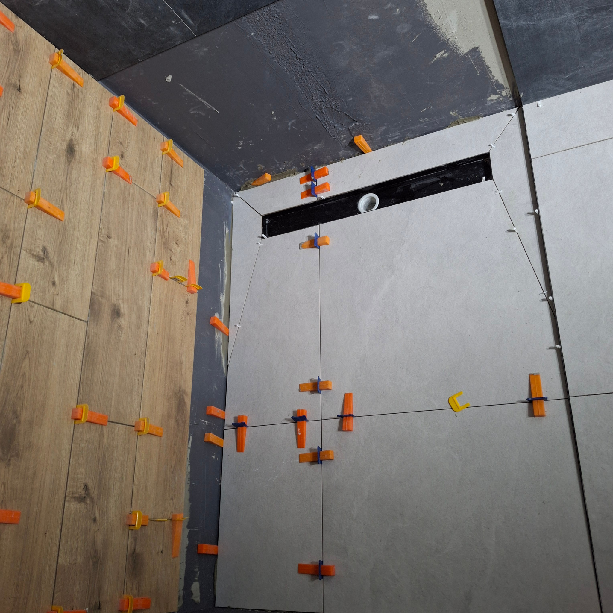 Układanie płytek drewnopodobnych i szarych w łazience z użyciem klipsów poziomujących i sznurka, widoczny odpływ liniowy w ścianie.