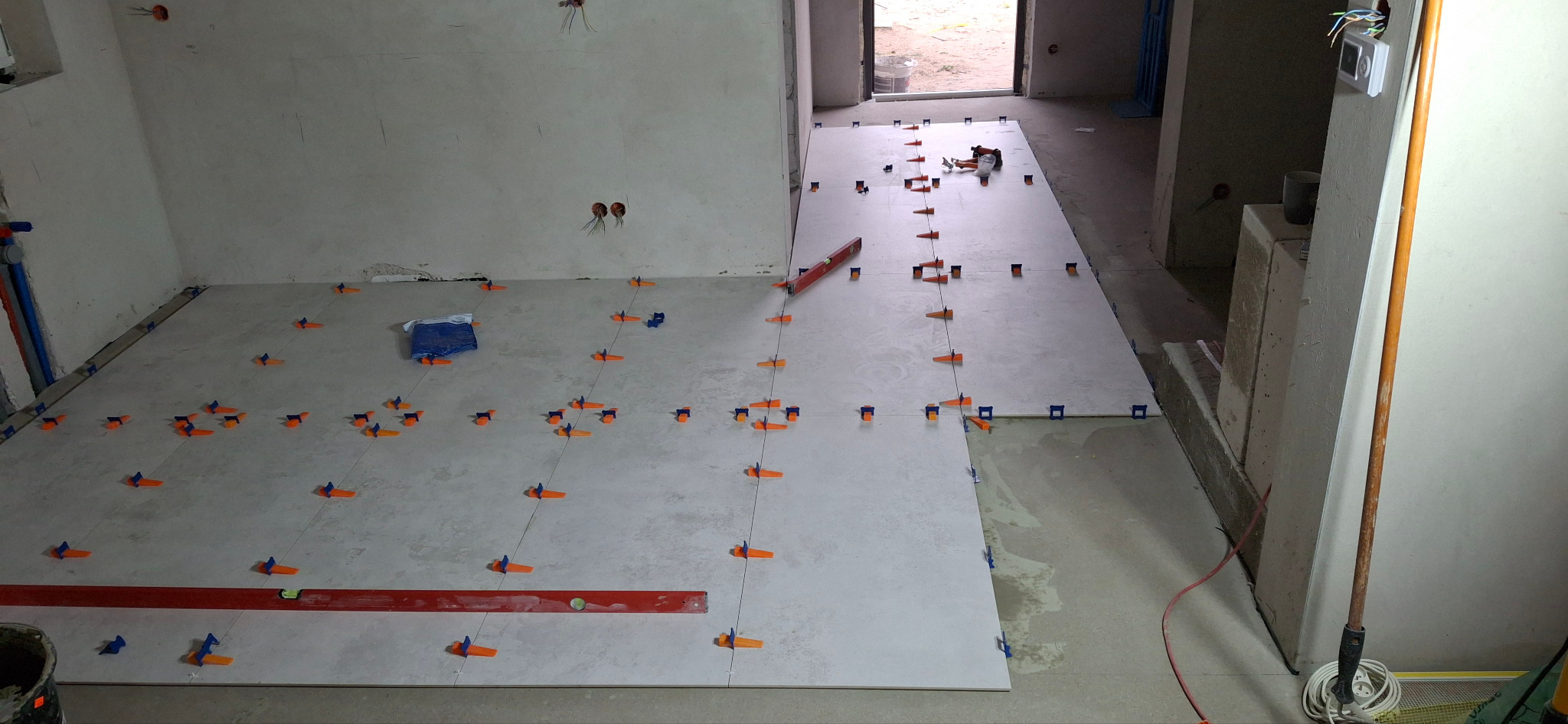 Układanie jasnych płytek podłogowych z użyciem poziomicy i pomarańczowych krzyżyków dystansowych w trakcie remontu łazienki, widoczne instalacje wodne i elektryczne.