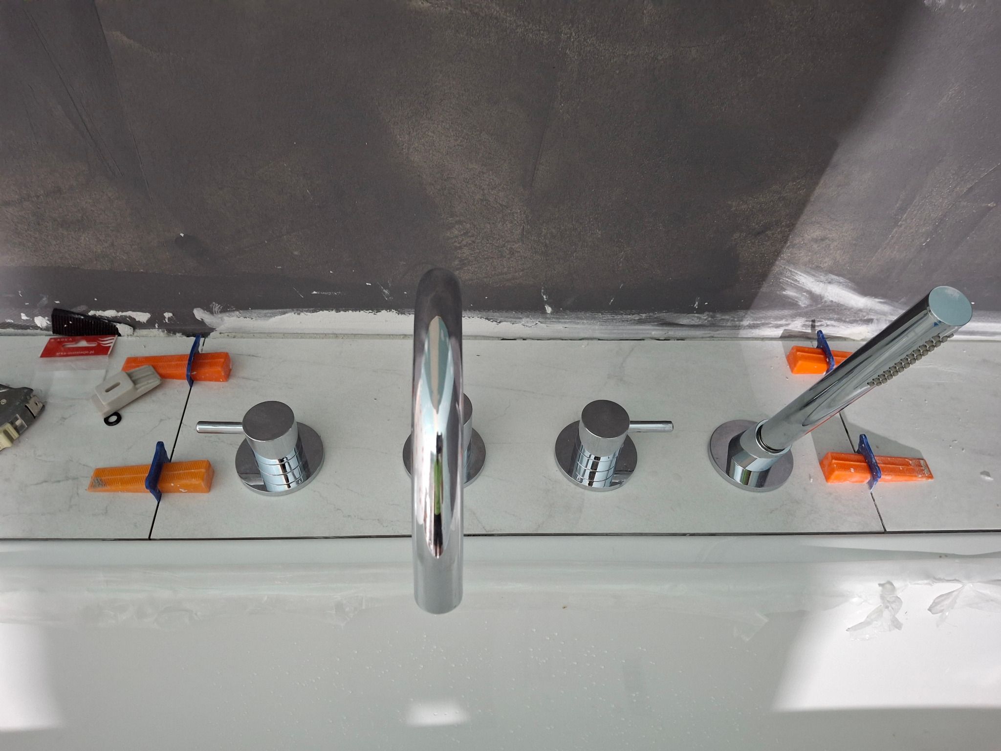 Bateria wannowa czterootworowa z chromowanymi pokrętłami i słuchawką prysznicową, zamontowana na białej wannie z obłożonymi płytkami ścianami, zabezpieczona pomarańczowymi klinami dystansowymi.