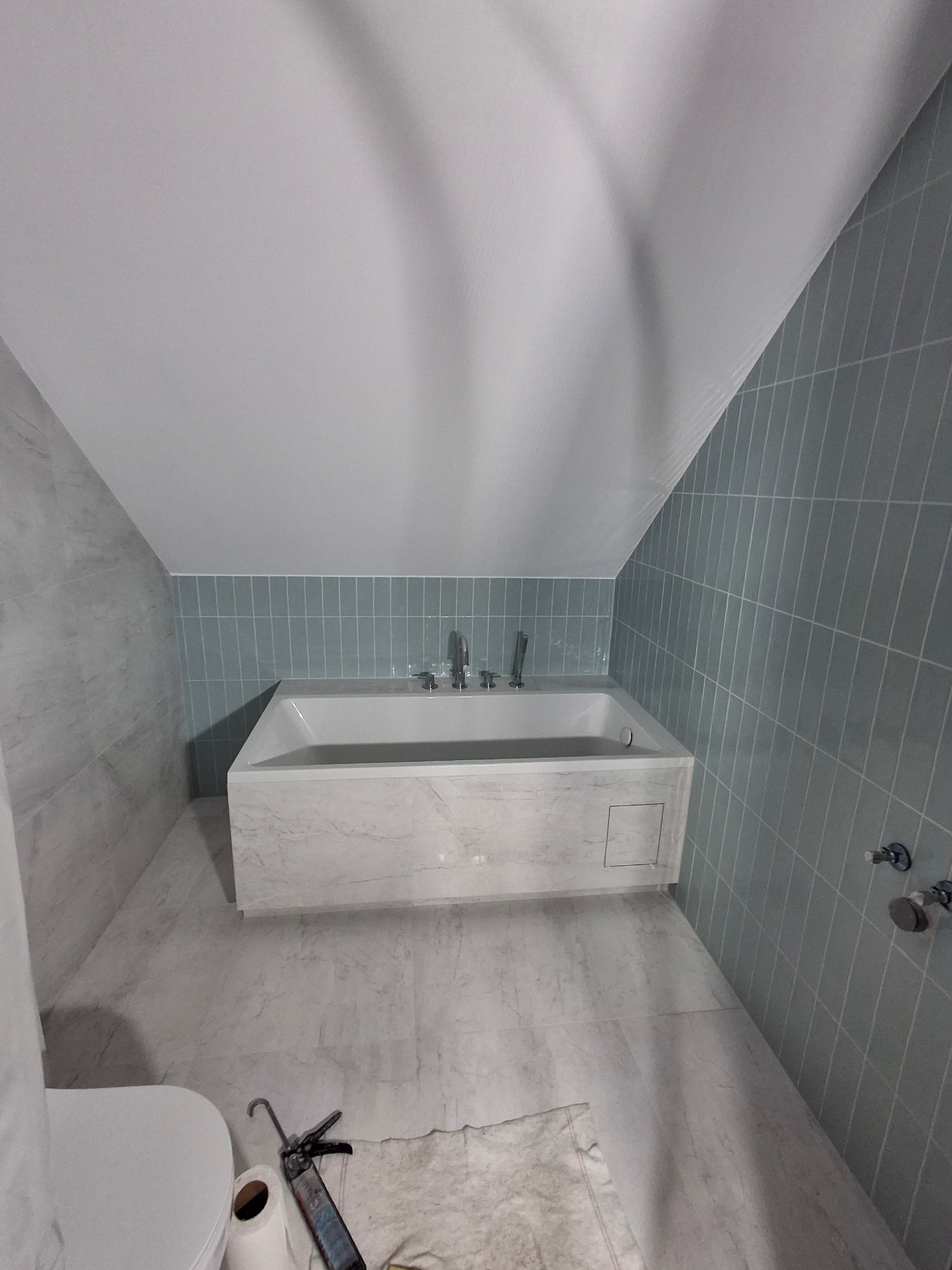 Wanna obudowana marmurem w łazience na poddaszu, ściany wyłożone pionowymi, błękitnymi płytkami, widoczne narzędzia i papier toaletowy na podłodze.