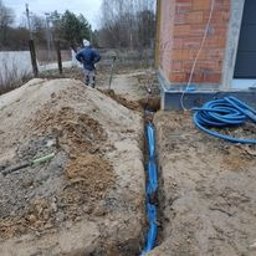 6EL - Wykopany rów z ułożonymi niebieskimi rurami kablowymi, obok hałda piasku i fragment budynku z cegły, pracownik w tle.