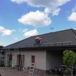 6EL - Pracownik w szortach montuje system odgromowy na dachu domu jednorodzinnego z czarną dachówką, widoczne rusztowania i drabiny, błękitne niebo z chmurami w tle.