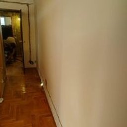 6EL - Świeżo pomalowana, biała ściana w korytarzu z parkietem. Widoczny fragment instalacji elektrycznej przy suficie oraz osoba pracująca w tle, w głębi korytarza.