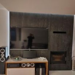 6EL - Ściana telewizyjna z szarego kamienia z podświetleniem LED, widoczny kominek i sprzęt audio, odbicie osoby w ekranie telewizora.