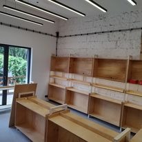 Surowy montaż instalacji elektrycznej w nowym lokalu handlowym: widoczne kable w czarnych rurkach na białej, ceglanej ścianie, surowe drewniane regały bez towaru, nowoczesne oświetlenie liniowe LED.