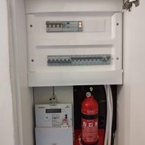 Otwarta szafka elektryczna z bezpiecznikami, licznikiem energii i gaśnicą proszkową w dolnej części.