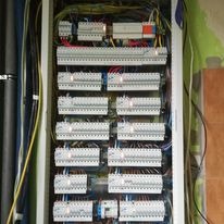 Otwarta skrzynka rozdzielcza z licznymi bezpiecznikami i przewodami elektrycznymi, widoczne elementy montażowe i okablowanie wewnątrz.