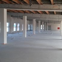 Surowe wnętrze hali w stanie deweloperskim z widocznymi słupami konstrukcyjnymi, betonową podłogą i odsłoniętym stropem z drewnianymi belkami. Na ścianie widoczny czerwony hydrant.