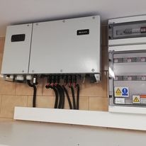 Szary inwerter fotowoltaiczny z podłączonymi kablami, zamontowany na białej półce, obok skrzynka elektryczna z naklejkami ostrzegawczymi na tle ceglanej ściany.