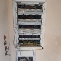 Otwarta skrzynka rozdzielcza z licznymi przewodami elektrycznymi, widoczne zabezpieczenia nadprądowe, kable wchodzące do skrzynki od dołu, ściana w tle z widocznymi puszkami instalacyjnymi.