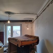 Niedokończona instalacja elektryczna w mieszkaniu, widoczne kable prowadzone po ścianie i suficie, prowizoryczne oświetlenie z żarówkami, w tle okno z widokiem na miasto, pomieszczenie w trakcie...