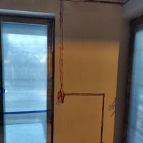 Ściana w trakcie remontu z wystającymi kablami elektrycznymi, widoczne puszki instalacyjne i przewody ułożone wzdłuż ściany, okno z roletami w tle.