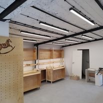 Wnętrze pomieszczenia w trakcie montażu oświetlenia: widoczne surowe, drewniane regały, lampy LED zamocowane na czarnych szynoprzewodach pod betonowym sufitem, częściowo wykończone ściany i podłoga.