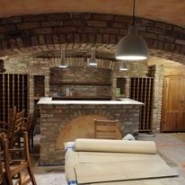 Piwnica z ceglanym sklepieniem łukowym, oświetlona trzema wiszącymi lampami. Widoczny ceglany bar z drewnianym blatem, regały na wino i zwój wykładziny podłogowej.