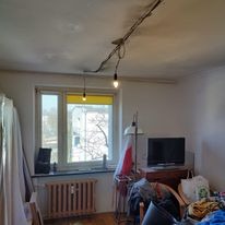 Niedokończona instalacja elektryczna w pokoju: widoczne przewody na suficie, prowizoryczne oświetlenie, grzejnik, okno z roletą, meble i przedmioty w nieładzie.