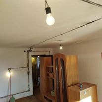 Wnętrze pokoju w trakcie remontu, widoczne prowizoryczne oświetlenie żarówkami na kablach, ściany z widocznymi przewodami elektrycznymi, meble odsunięte od ścian.