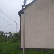 Słup oświetleniowy z kamerą monitoringu zamontowany przy ścianie budynku w Częstochowie. Widoczne okablowanie i trawnik.
