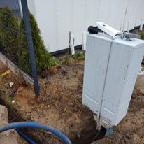 Biała skrzynka elektryczna wystająca z ziemi w wykopie, widoczne kable i narzędzia, tło z krzewami i ścianą budynku.