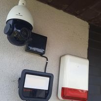 Zewnętrzna kamera obrotowa PTZ z reflektorem i sygnalizatorem alarmowym zamontowana na jasnej ścianie budynku, widoczne okablowanie.