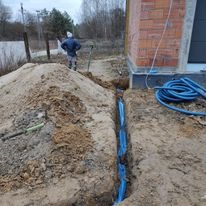 Wykopany rów z ułożonymi niebieskimi rurami kablowymi, obok hałda piasku i fragment budynku z cegły, pracownik w tle.