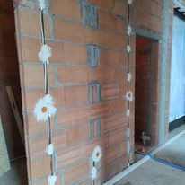 Ściana z cegły ceramicznej z pionowo poprowadzonymi przewodami elektrycznymi, zabezpieczonymi białym gipsem w miejscach mocowania, widoczne puszki instalacyjne w ścianie.