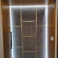 Panel wejściowy z szarego materiału, obramowany drewnem i podświetlony ledowym oświetleniem po bokach, z czarną klamką po lewej stronie i panelem sterowania po prawej.