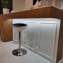 Podświetlona diodami LED biała zabudowa meblowa pod blatem z drewnopodobną okleiną, widoczny metalowy hoker barowy na tle jasnej podłogi.