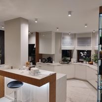 Nowoczesna, jasna kuchnia z białymi szafkami i marmurowym blatem, oświetlona punktowymi lampami sufitowymi. Widoczny fragment barku z podświetleniem LED i hokera.