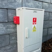 Biała skrzynka elektryczna z ostrzeżeniami na tle szarej ściany z betonowych bloczków. Na skrzynce widoczne naklejki ostrzegawcze o wysokim napięciu i zagrożeniu porażeniem. Dodatkowo czerwona...