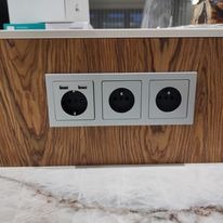 Potrójne gniazdo elektryczne z dwoma portami USB, zamontowane w obudowie z drewna o wyraźnym usłojeniu, widoczne na tle marmurowego blatu.